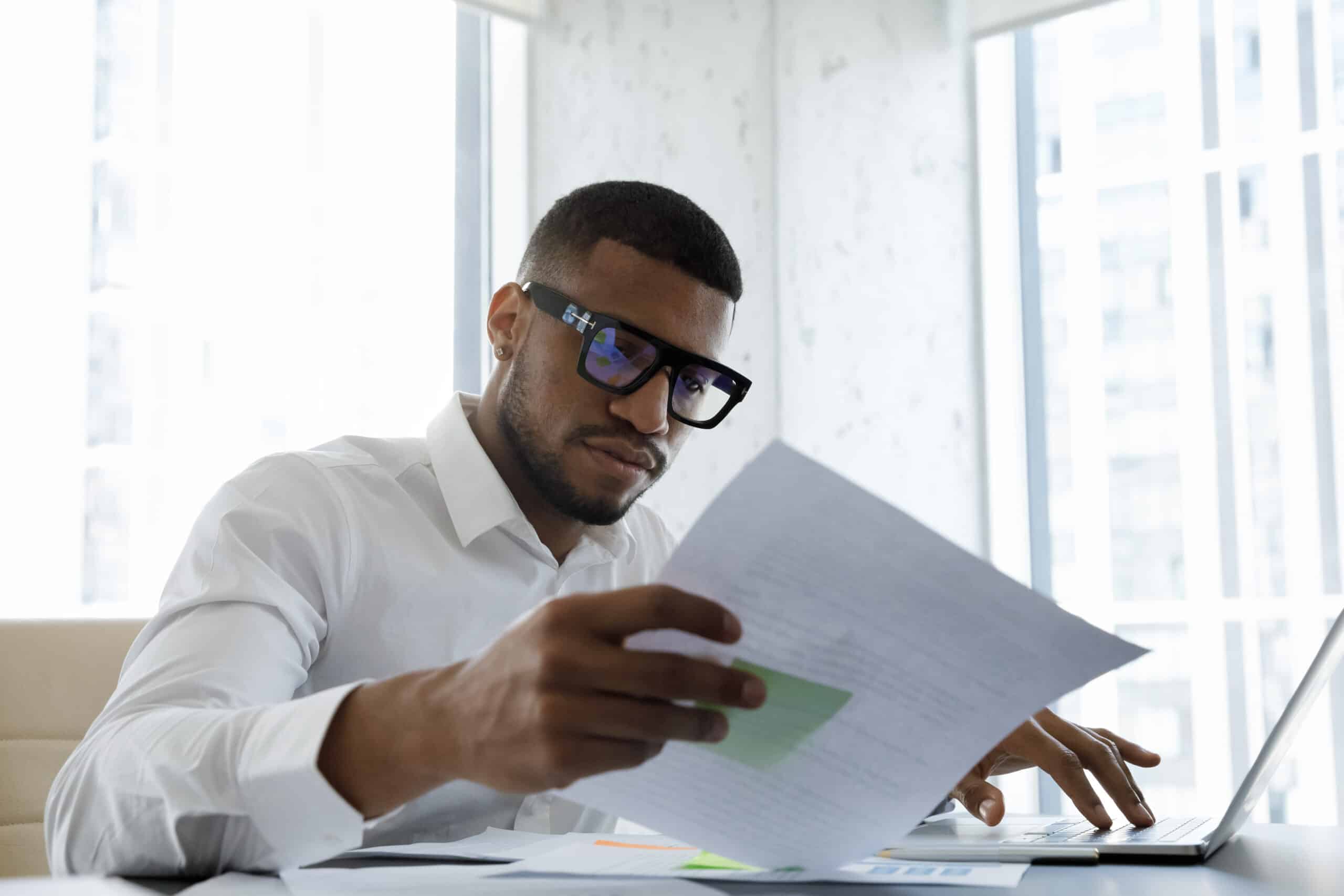 Serious African American manager in stylish glasses reading corporate document with note, sales report, boss order, doing paperwork. Legal expert checking document at workplace with laptop