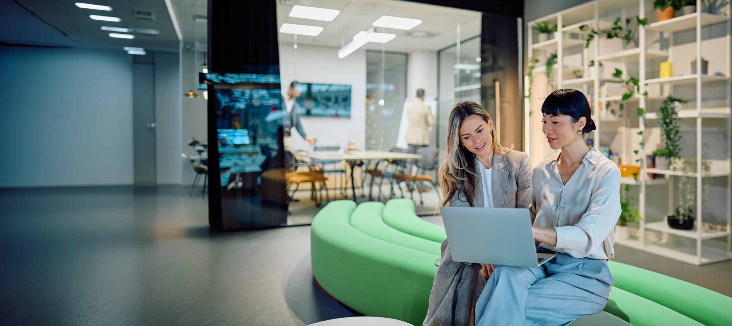 Two coworkers sitting on a green bench, working together on a laptop.