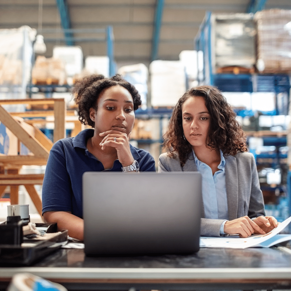 Two women working at a laptop in a warehouse.